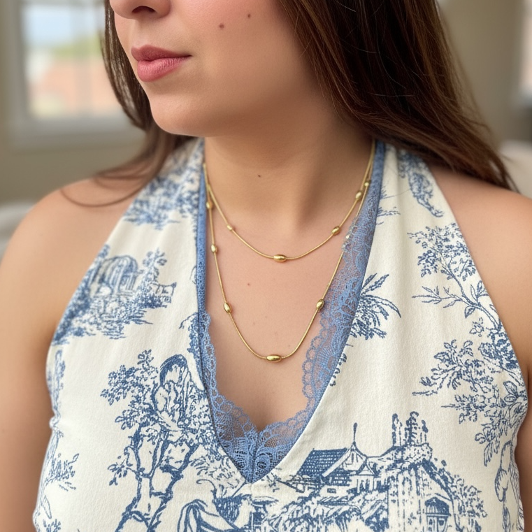 Woman wearing a blue and white patterned top with gold necklaces indoors.