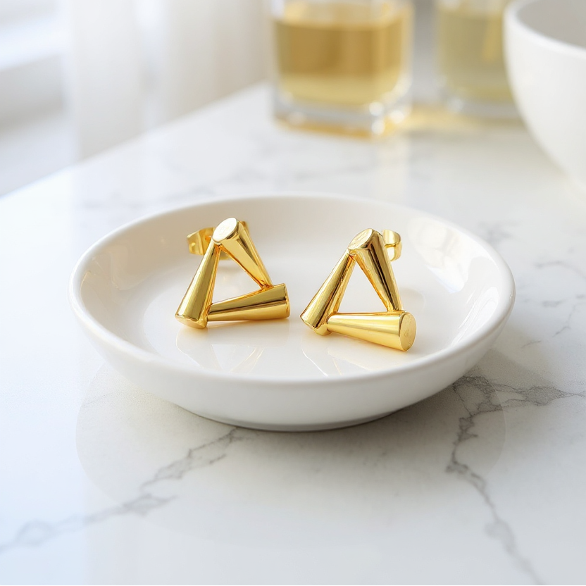Gold triangle-shaped earrings on a white dish with a blurred background of bottles.